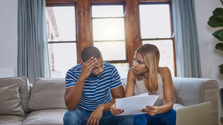 Man and woman sit on a living room couch examining papers; the man looks stressed as the woman explains the documents.