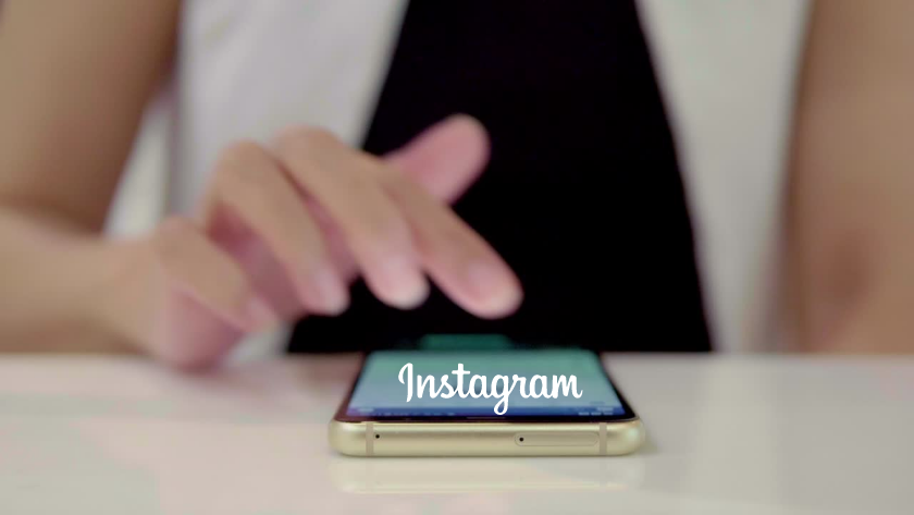 Person's finger taps a smartphone displaying the Instagram logo on a reflective table surface.