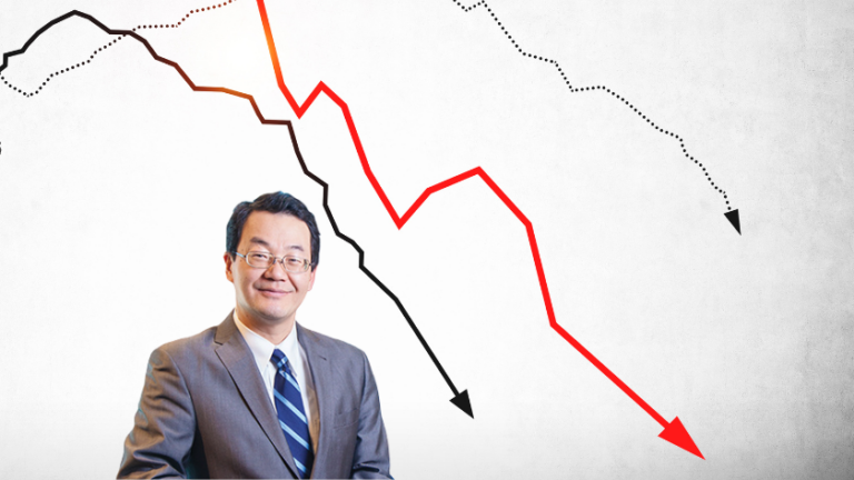 Businessman in a suit stands in front of downward-trending stock charts with red and black lines and arrows indicating decline.