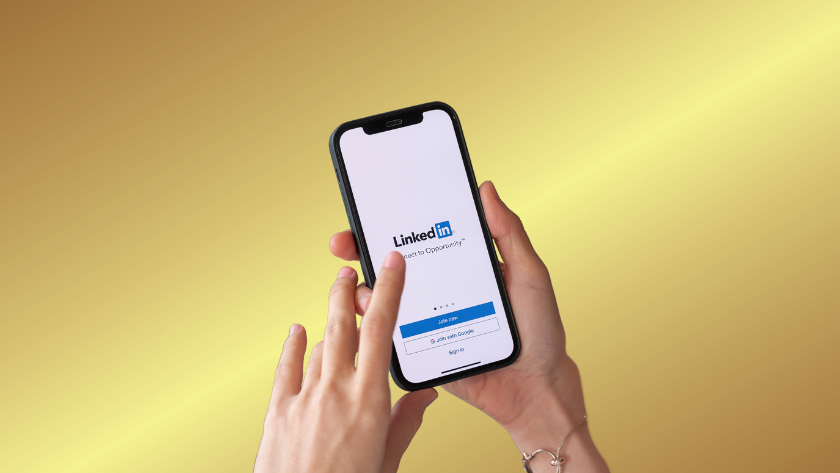 Hand holding a smartphone displaying the LinkedIn login screen against a yellow gradient background.