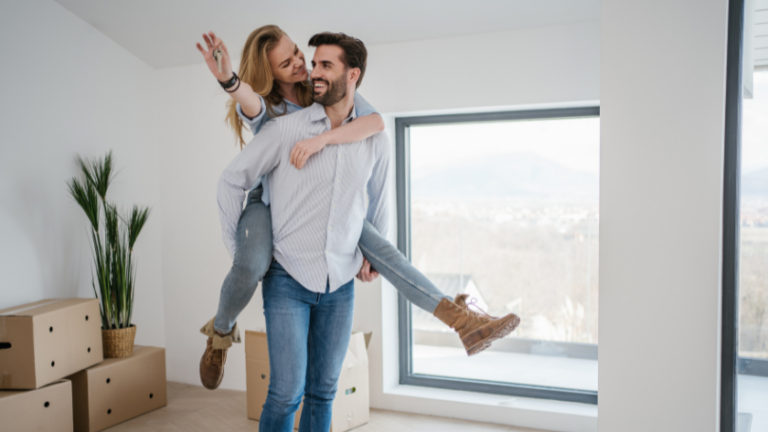 Couple in a bright living room, man carrying a woman on his shoulders, surrounded by moving boxes and a large window view behind them.