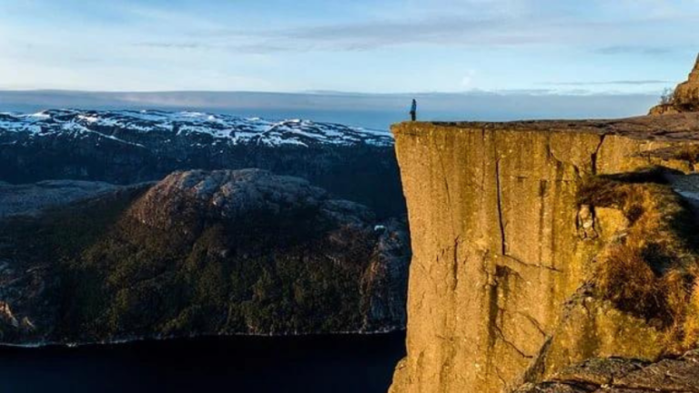 Person standing at the edge of a tall cliff, overlooking a dark fjord and snow-capped mountains in the distance at sunset or morning light.