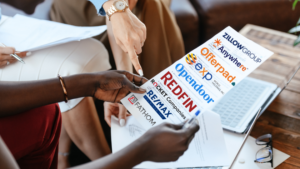 Group of diverse hands sharing a brochure featuring real estate company logos (Redfin, OpenDoor, Offerpad, Zillow Group, Exto, RE/MAX, Fathom) on a table with a laptop and documents nearby.