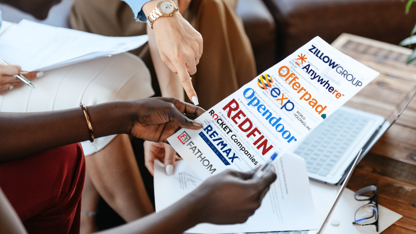 Group of diverse hands sharing a brochure featuring real estate company logos (Redfin, OpenDoor, Offerpad, Zillow Group, Exto, RE/MAX, Fathom) on a table with a laptop and documents nearby.