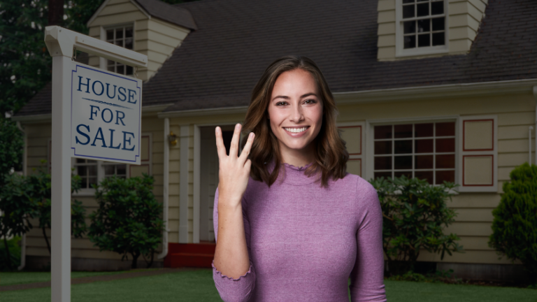 Smiling woman in a pink sweater stands in front of a beige house with a 'House for Sale' sign in the yard
