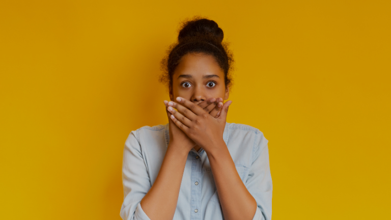 Woman with a surprised expression covers her mouth with both hands against a solid mustard yellow background, wearing a light denim shirt.