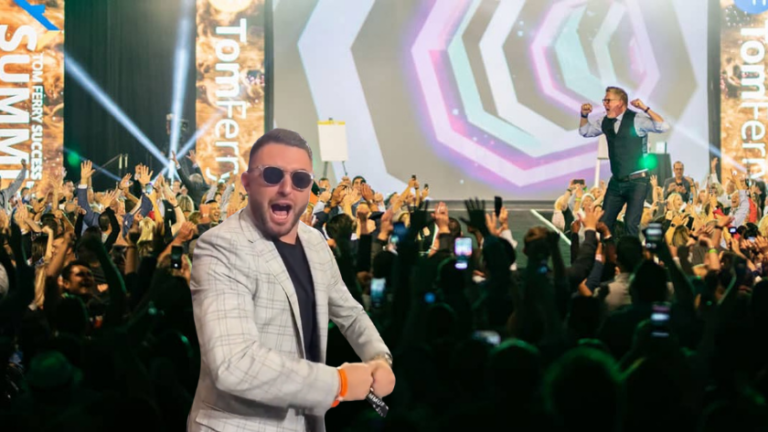 Man in a light plaid blazer and sunglasses dances at a packed concert, microphone in hand, with colorful stage lights behind him.