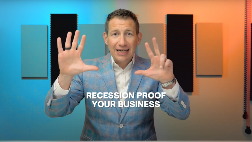 Man in a light blue blazer speaks to camera with hands raised, the text 'Recession Proof Your Business' overlaid in white. Background shows sound panels and colorful lighting.