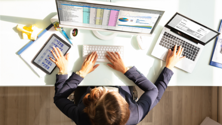 Top-down view of a person at a glass desk typing on a keyboard with a tablet, a laptop, and two monitors displaying charts and spreadsheets.