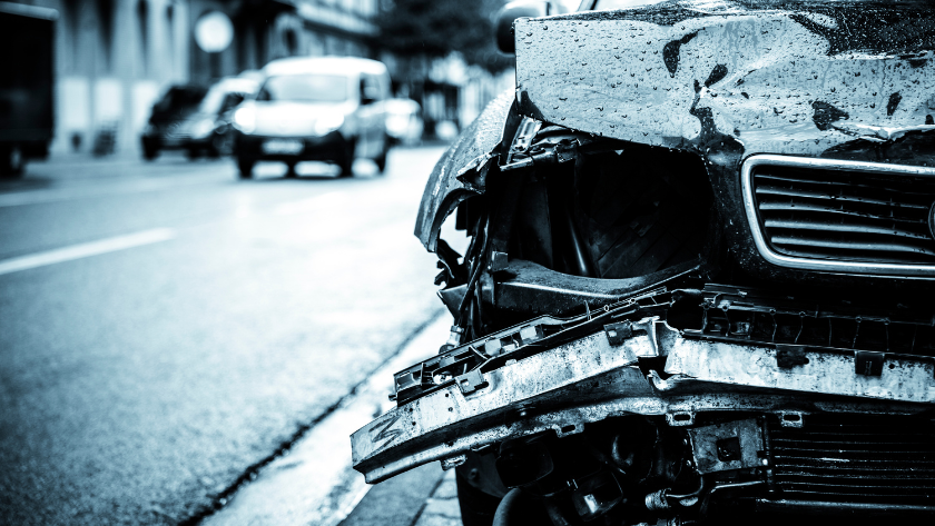 Front of a damaged car with a crumpled hood and exposed components on a city street after a collision.