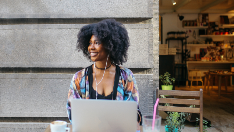 Woman with curly hair working on a laptop at an outdoor cafe, wearing a colorful cardigan and white earphones, smiling.
