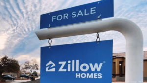 Blue 'For Sale' sign with a Zillow Homes placard hanging beneath, in a residential street scene on a clear day.