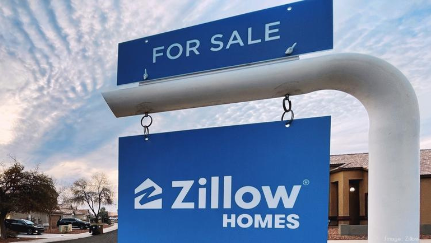Blue 'For Sale' sign with a Zillow Homes placard hanging beneath, in a residential street scene on a clear day.