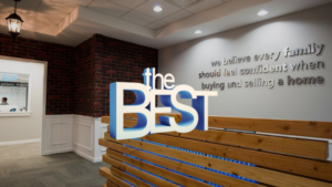Lobby of a real estate office with a large 3D 'the BEST' sign, brick accent wall, and a wooden slat bench under warm lighting.