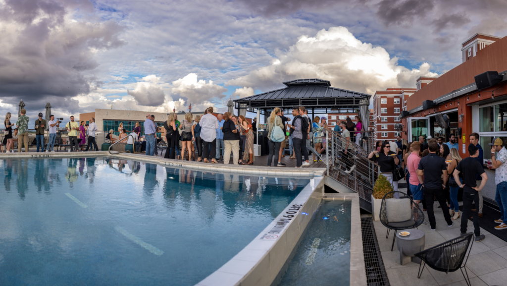 Crowd of adults mingling by a rooftop pool under cloudy skies, with a central gazebo and surrounding brick buildings in the background.