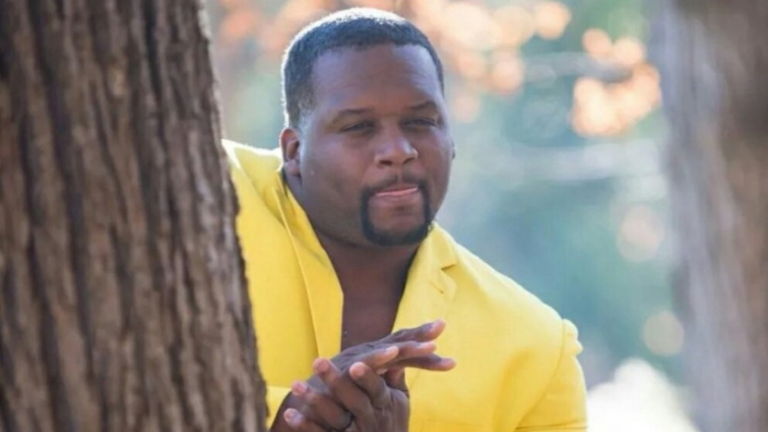Man in a bright yellow blazer leaning against a tree outdoors, looking to the side.