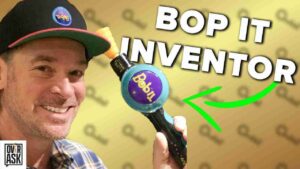 Smiling man in a cap holds a Bop It toy; bold 'BOP IT INVENTOR' text and a neon green arrow highlight the device.