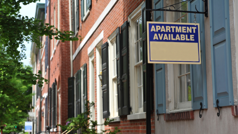 Blue and yellow 'Apartment Available' sign hanging from a black bracket on a brick row house with blue shutters and greenery nearby.