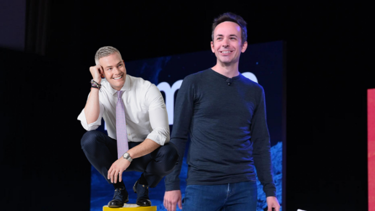 Two men on a stage smiling; one crouches on a yellow block in a white shirt and tie, the other stands beside him in a dark long-sleeve shirt.