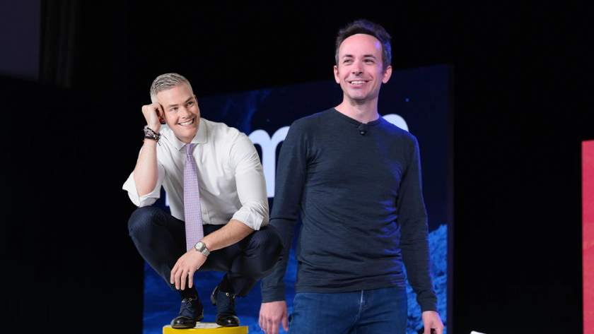 Two men on a stage smiling; one crouches on a yellow block in a white shirt and tie, the other stands beside him in a dark long-sleeve shirt.