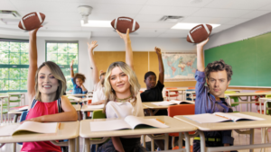 Diverse students in a bright classroom raise their hands, each holding a football, seated at desks with notebooks open.