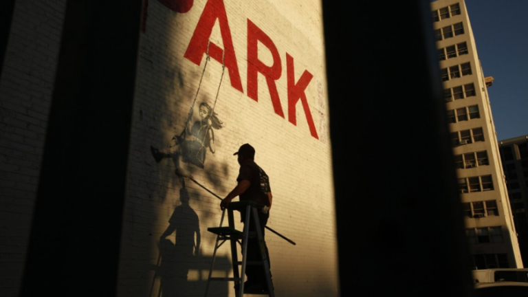 Worker on a ladder painting a mural on a brick wall, with the large red word 'PARK' and shadows on the ground.