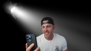 Man in a white t-shirt and backward cap holds a smartphone, filming a selfie under a bright stage spotlight.