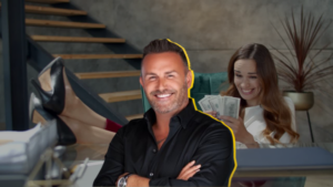 Smiling man in a black shirt stands in the foreground as a woman in the background counts cash in an office setting.