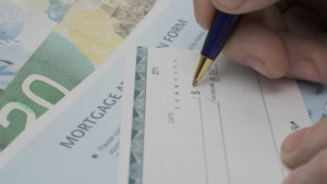Close-up of a hand writing on a mortgage check form with a blue pen and cash in the background.