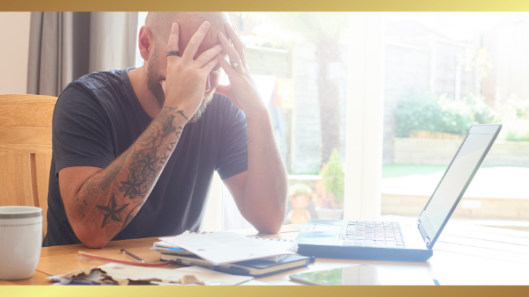 Bearded man with tattoos sits at a table with his head in his hands, looking stressed at an open laptop and scattered papers in a bright room.