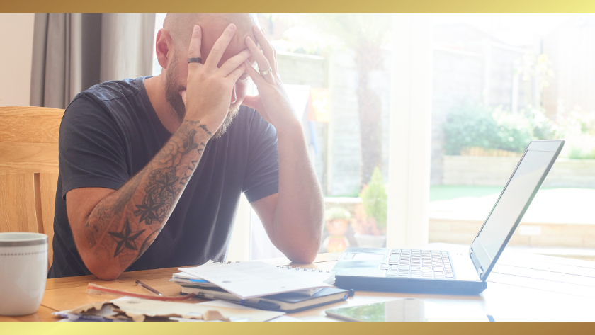 Bearded man with tattoos sits at a table with his head in his hands, looking stressed at an open laptop and scattered papers in a bright room.