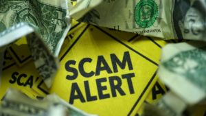 Bright yellow sign reads SCAM ALERT surrounded by crumpled dollar bills and torn paper.