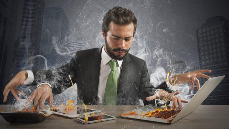 Businessman in a suit multitasks across a laptop, tablet, and phone as flames rise around him
