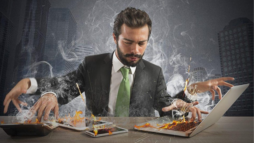 Businessman in a suit multitasks across a laptop, tablet, and phone as flames rise around him