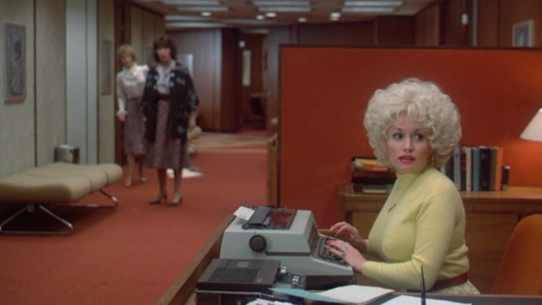 Woman with a voluminous 1960s hairstyle sits at a desk typing on a vintage computer while two women walk toward the hallway behind her in a retro office setting.