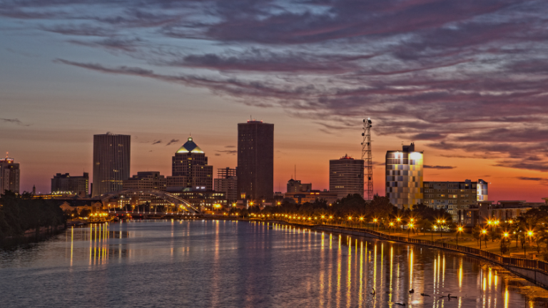 City skyline at sunset with a river in the foreground and glowing streetlights reflecting on the water.