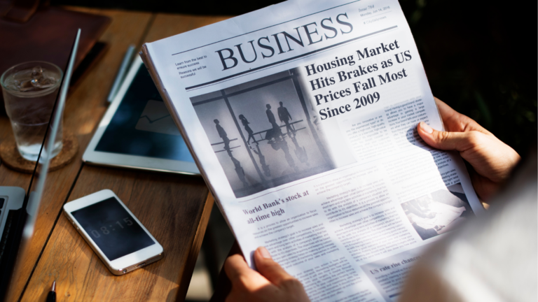 Person reads a newspaper on business news about housing prices, with a glass, tablet, and smartphone on the wooden table nearby.
