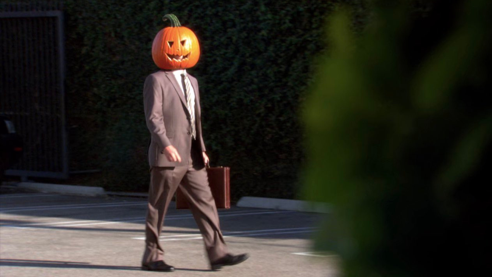 Person in a brown business suit walking in a parking lot with a carved pumpkin head and carrying a briefcase in Halloween attire.