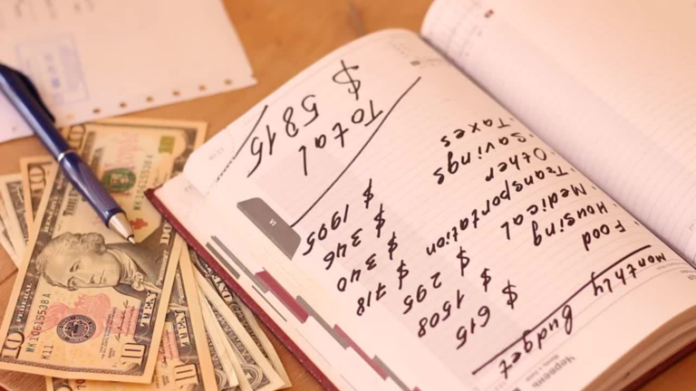 Notebook page with handwritten financial numbers and a dollar sign, surrounded by scattered ten-dollar bills and a blue pen on a desk.