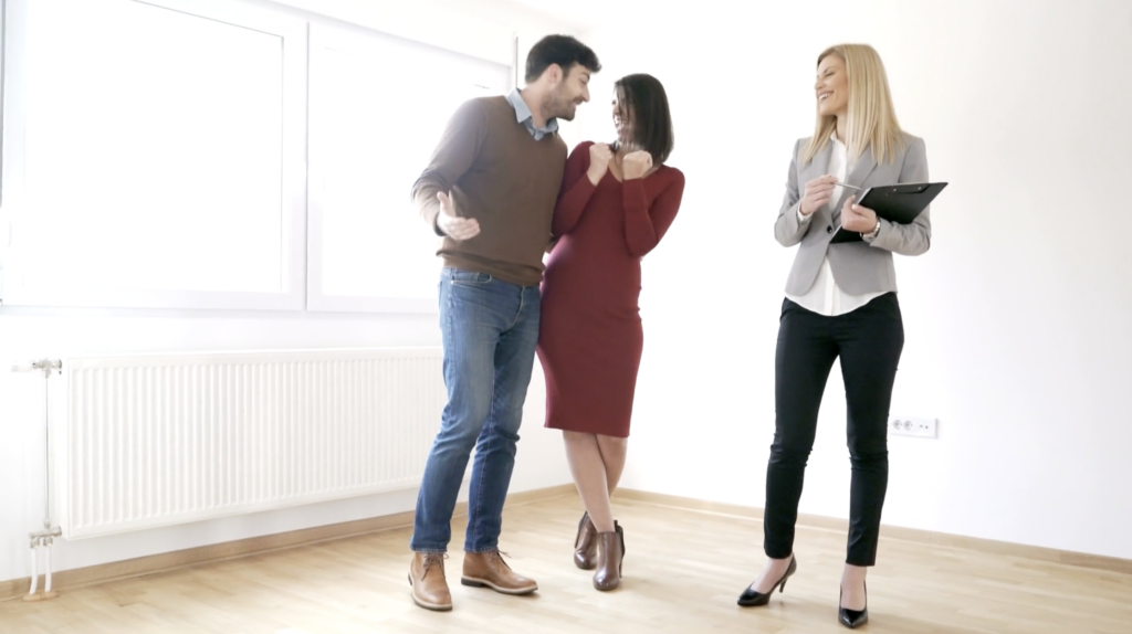 Three colleagues in a bright room chatting and smiling together, a man left, a woman in red in the middle, and a woman with a clipboard on the right.