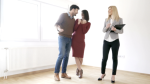 Three colleagues in a bright room chatting and smiling together, a man left, a woman in red in the middle, and a woman with a clipboard on the right.