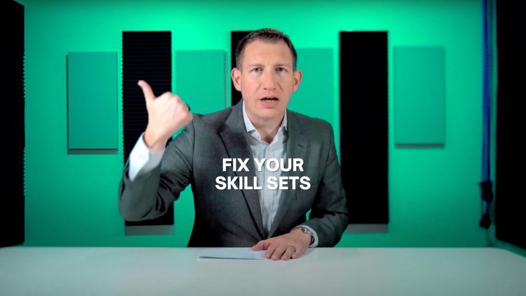 Man in a gray suit sits at a white desk in a green studio, gesturing with his left hand while a note rests on the desk; overlay reads 'FIX YOUR SKILL SETS'.