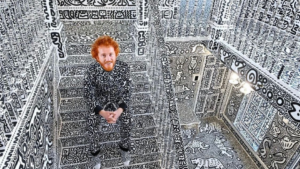 Man with red hair and beard sits in a room whose walls, floor, and ceiling are covered in black-and-white doodle drawings.