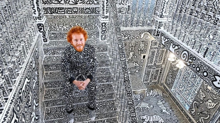 Man with red hair and beard sits in a room whose walls, floor, and ceiling are covered in black-and-white doodle drawings.