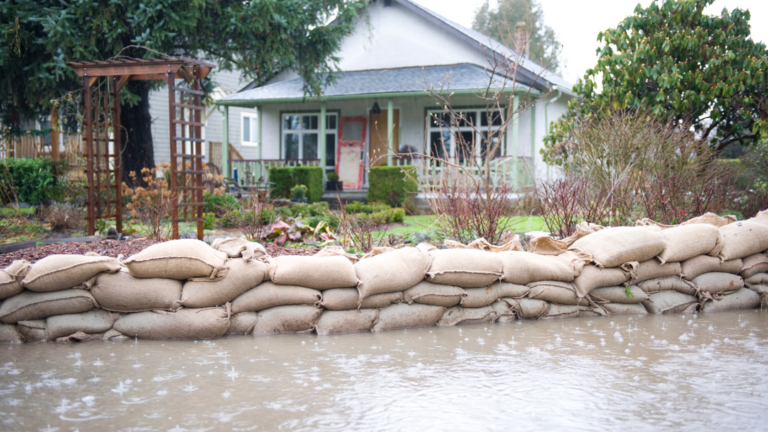 Flood barrier of stacked burlap sandbags along a flooded yard in front of a house with a garden and trellis.