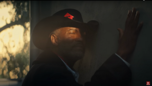 Man wearing a black cowboy hat with a red emblem leans against a wall, eyes closed, bathed in warm sunlight.