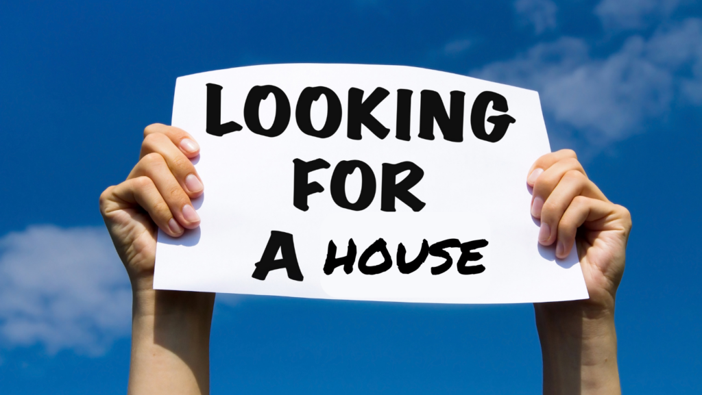 Person holds a white sign that says “LOOKING FOR A HOUSE” against a clear blue sky.