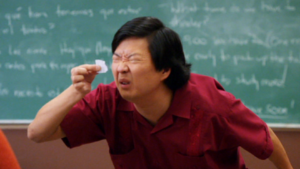 Man in a red shirt squints and holds a crumpled tissue to his eye in a classroom with a chalkboard in the background.