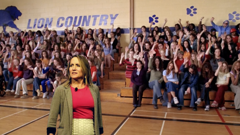 Woman in pink top and gray blazer speaks to a large gymnasium audience of students with hands raised, 'LION COUNTRY' mural in the background.