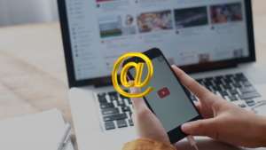 Person holds a smartphone showing the YouTube app logo with a large gold @ symbol floating in front.